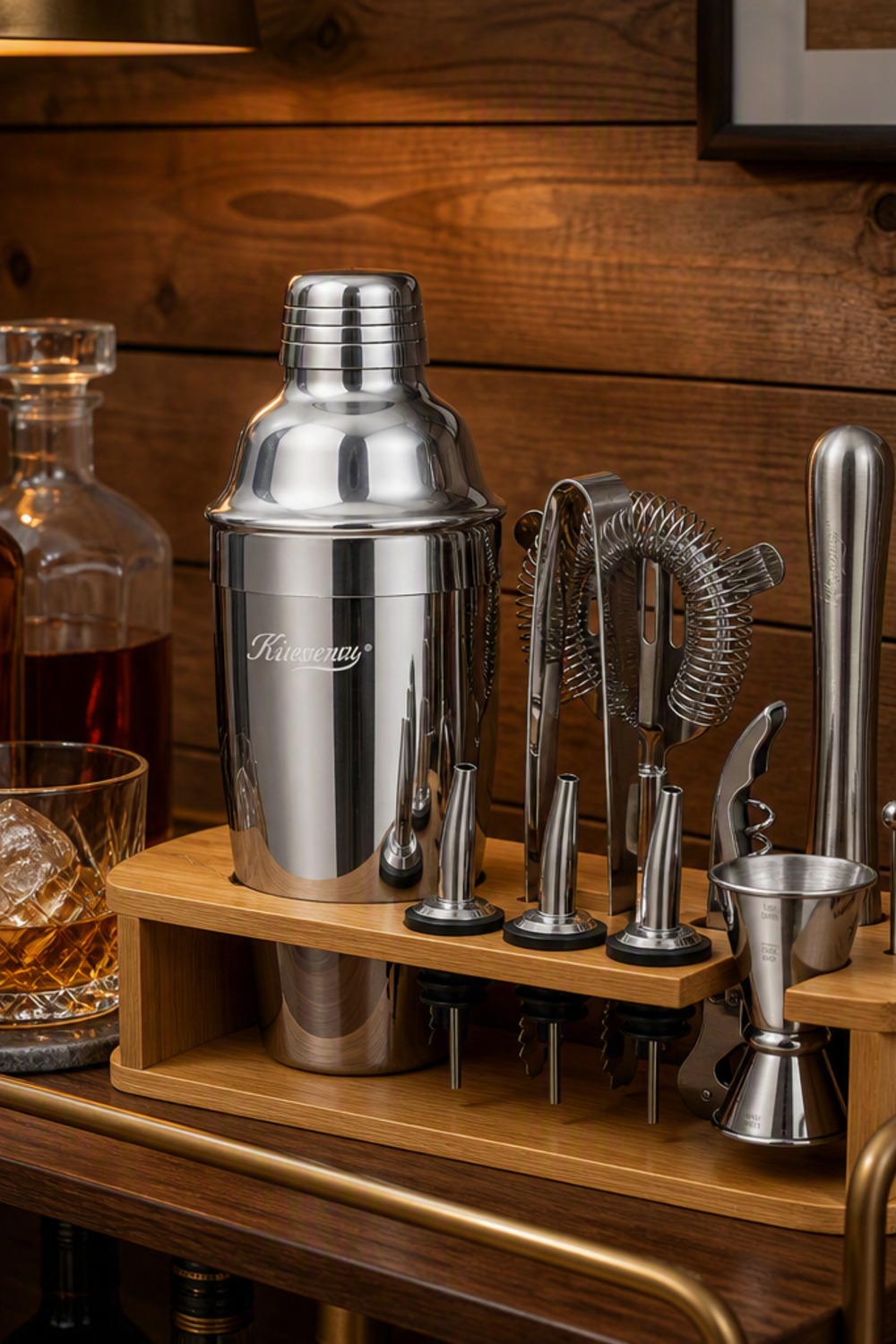 Stainless cocktail shaker set with jigger, strainer, and muddler on a bamboo stand on a cabin bar cart