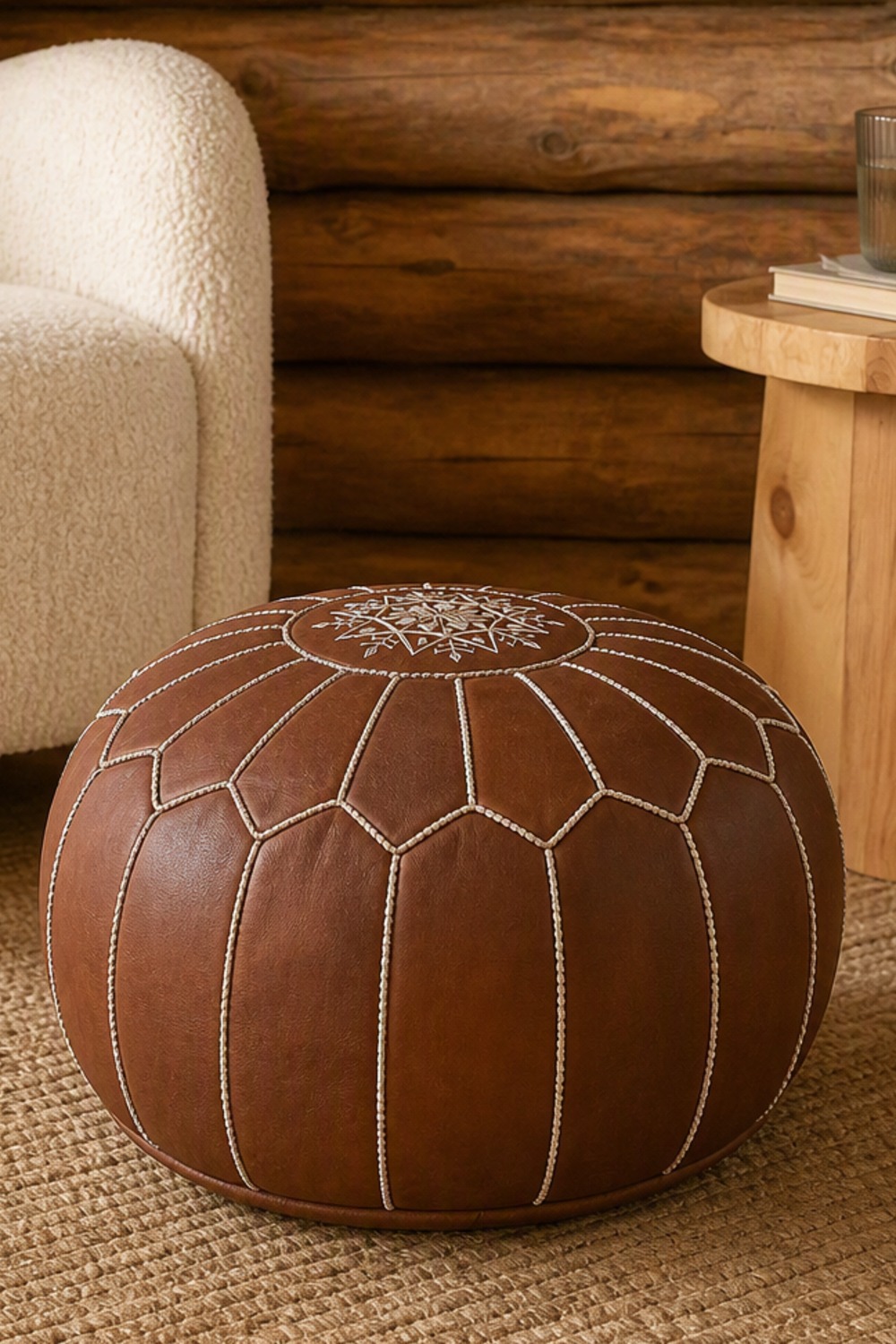 Round cognac-brown leather Moroccan pouf with hand-stitched panels and an embroidered star medallion, on a woven rug beside a cream boucle armchair