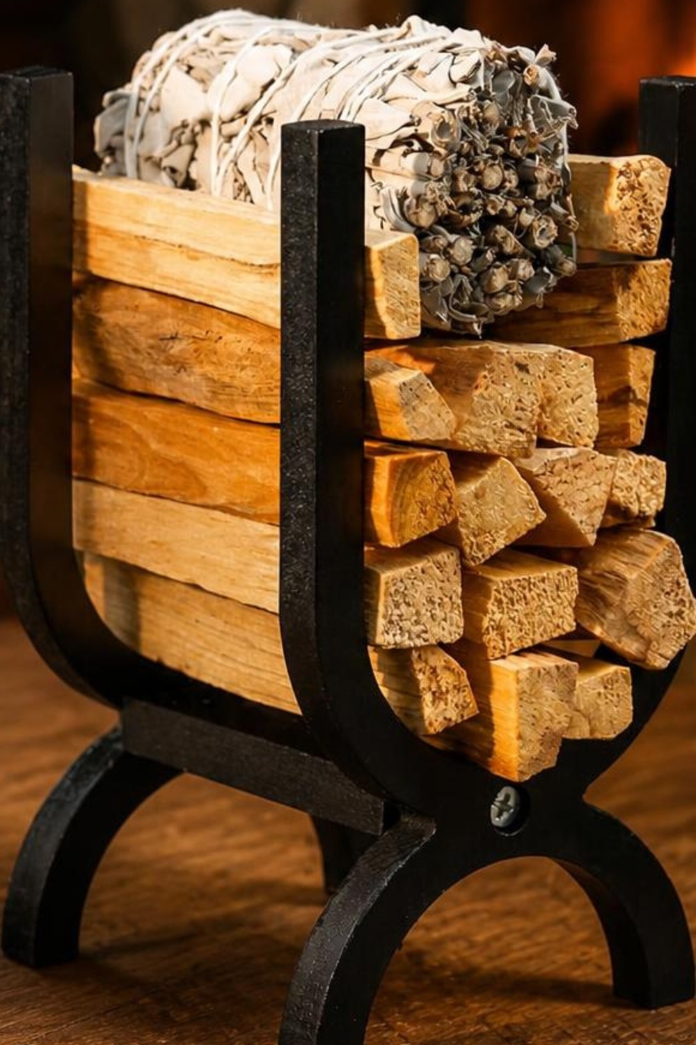Small black U-shaped wooden firewood and palo santo holder stacked with wood chunks and a white sage smudge bundle