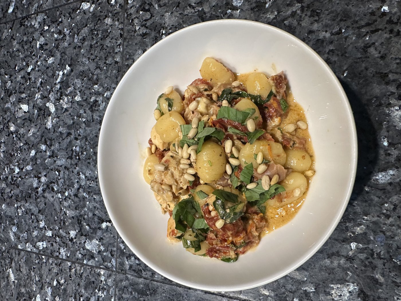 A bowl of chicken gnocchi with gorgonzola, sun-dried tomatoes, prosciutto, pine nuts, and fresh basil on a granite counter
