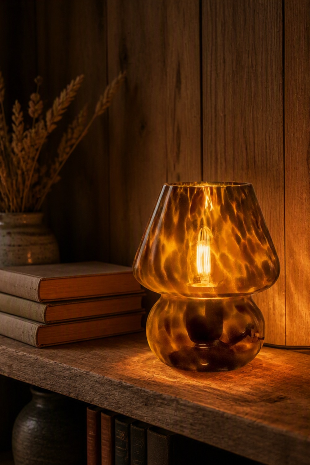 Tortoise-shell glass mushroom table lamp glowing amber on a rustic wood shelf beside vintage books and dried grasses in a cabin