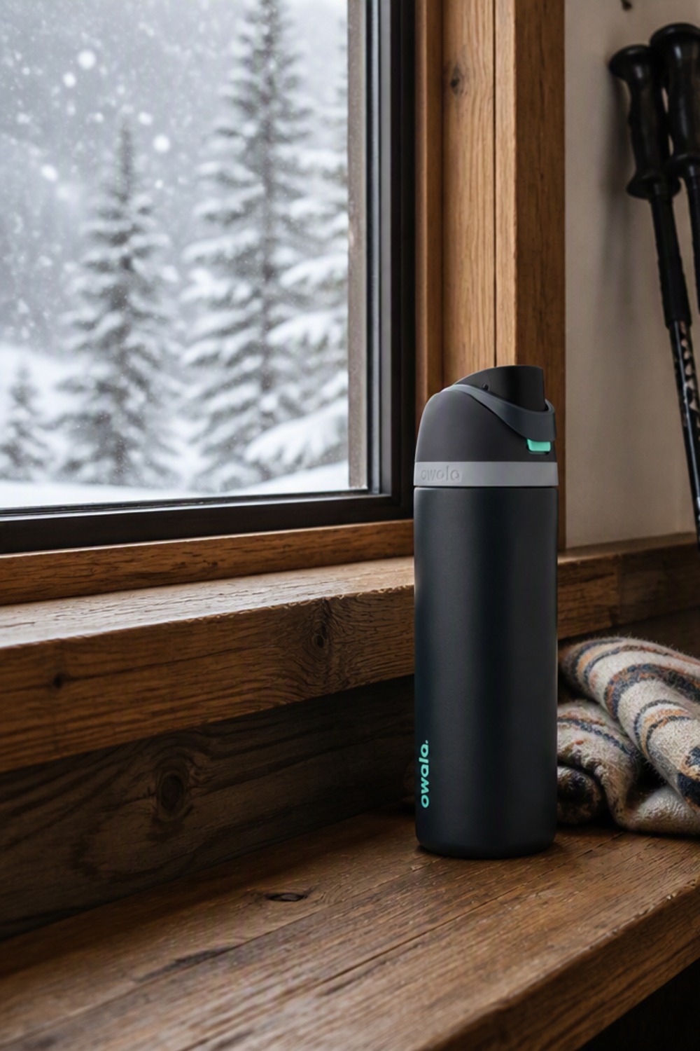 Matte black Owala FreeSip insulated water bottle with mint-green accents on a rustic cabin windowsill, ski poles and snow visible outside