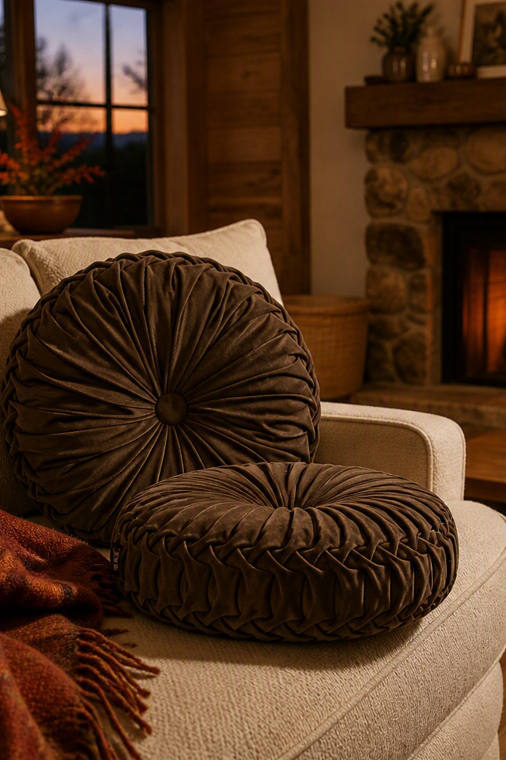 Two chocolate brown round pleated velvet pillows on an oatmeal sofa by a stone fireplace at dusk