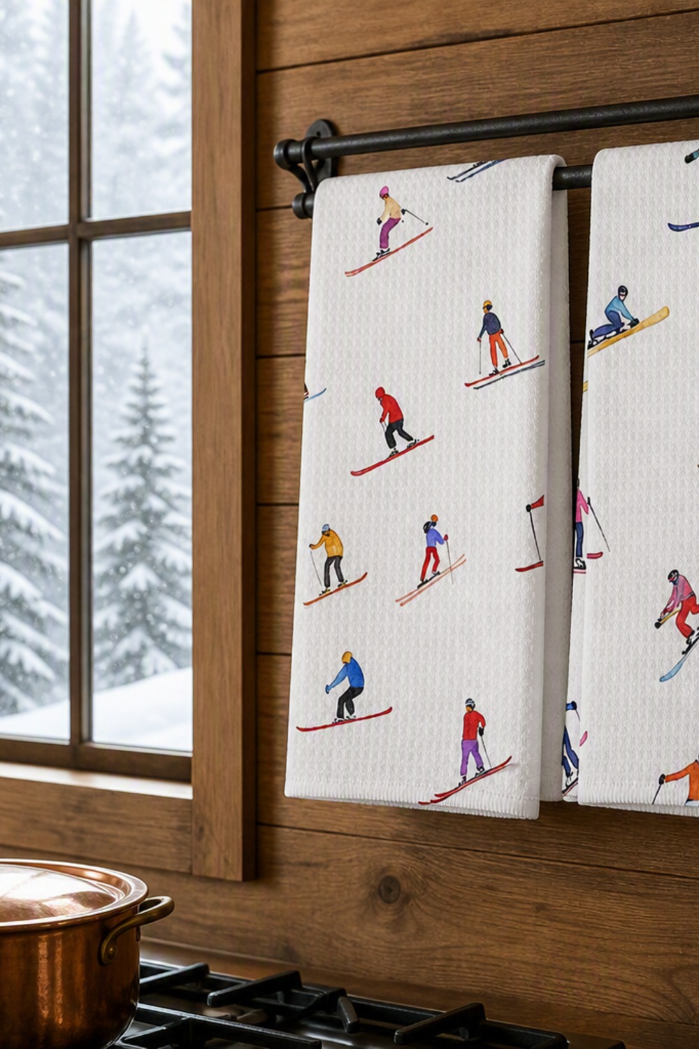 Two kitchen towels printed with colorful illustrated skiers and snowboarders, hanging beside a snowy window in a cabin kitchen
