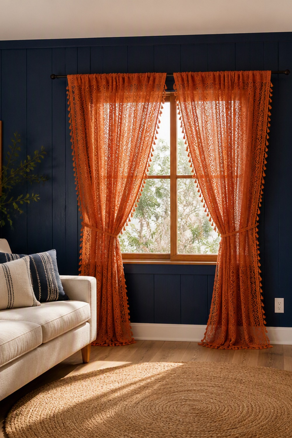 A pair of sheer burnt-orange crochet-lace curtain panels with tassel trim, tied back beside a wood-framed window against deep navy board-and-batten walls, above a cream linen sofa and a round jute rug