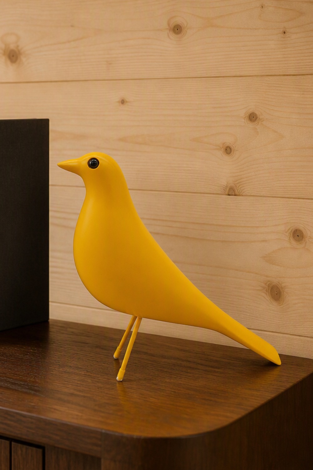 Mustard-yellow mid-century-style bird sculpture on thin yellow legs, standing on a dark walnut console beside a standing book against a pine-plank wall